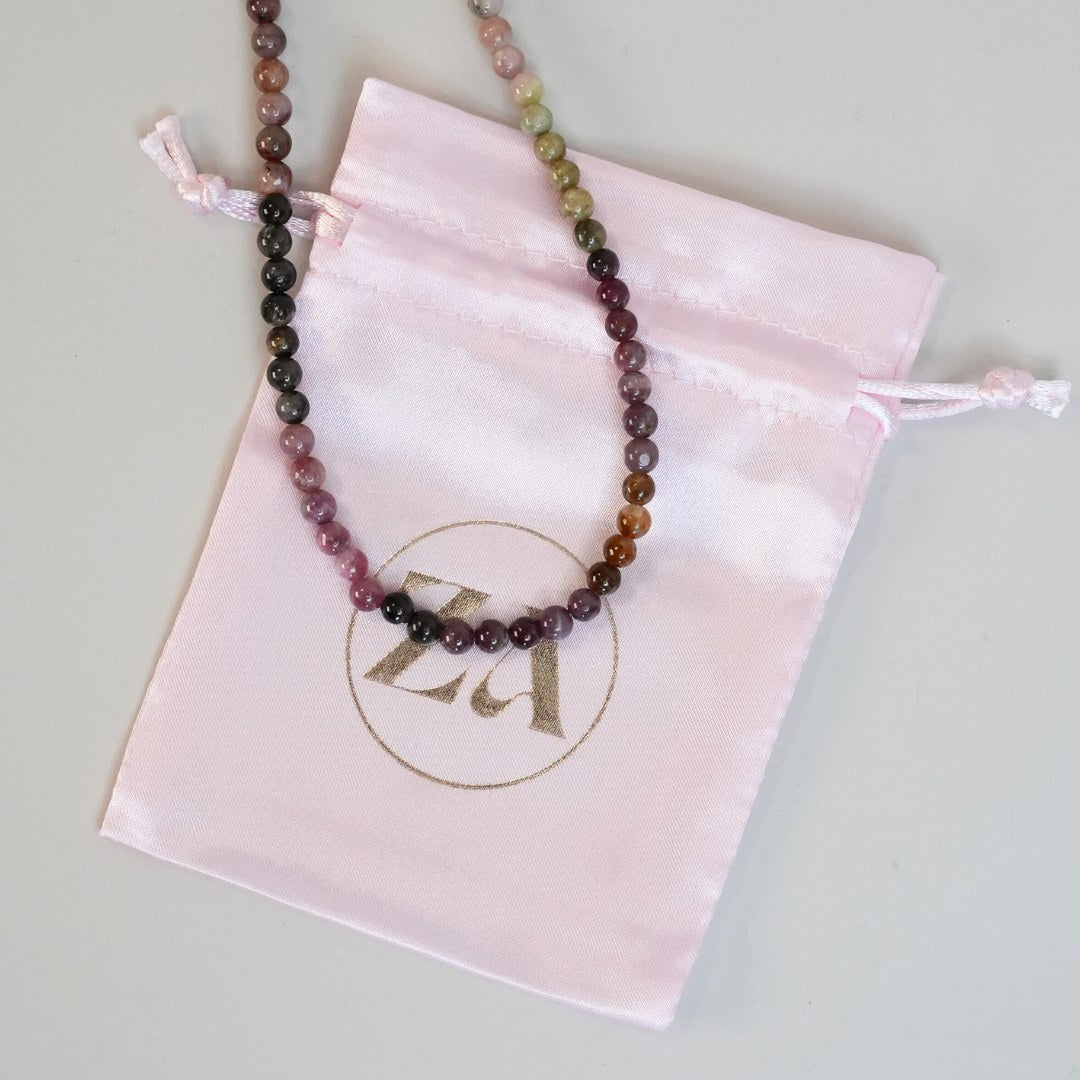 Et farverigt halskæde med perler i forskellige nuancer af lilla, grøn og brun ligger på en lyserød silkepose med en guldfarvet logo ZÆ.