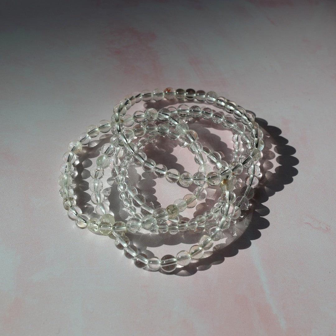 Billede af flere runde armbånd lavet af bjergkrystal med gennemsigtige perler liggende på en lys pink overflade med skygger