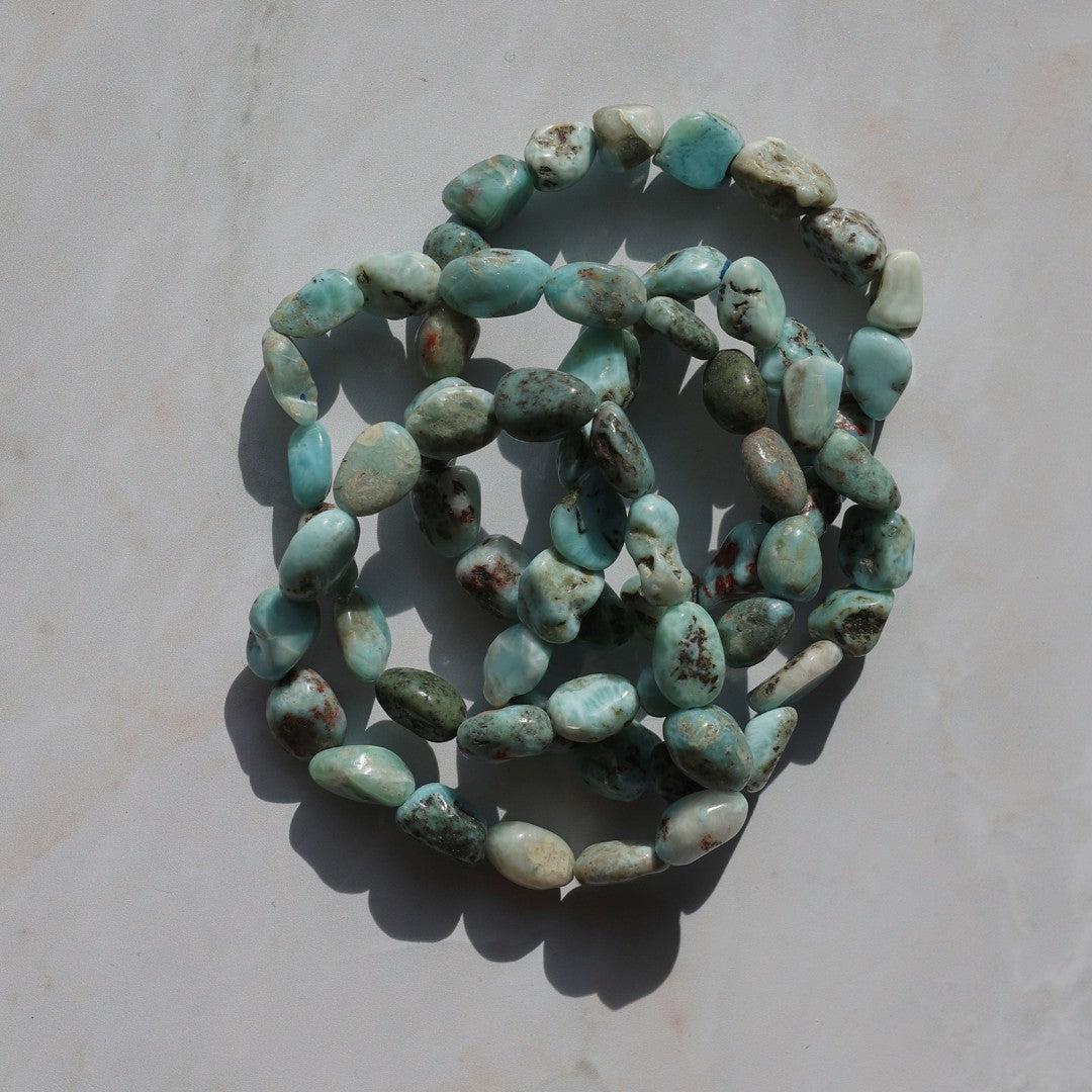 Larimar armbånd lavet af slebne sten i nuancer af blå, grøn og grå samlet i en spiralform på en lys baggrund