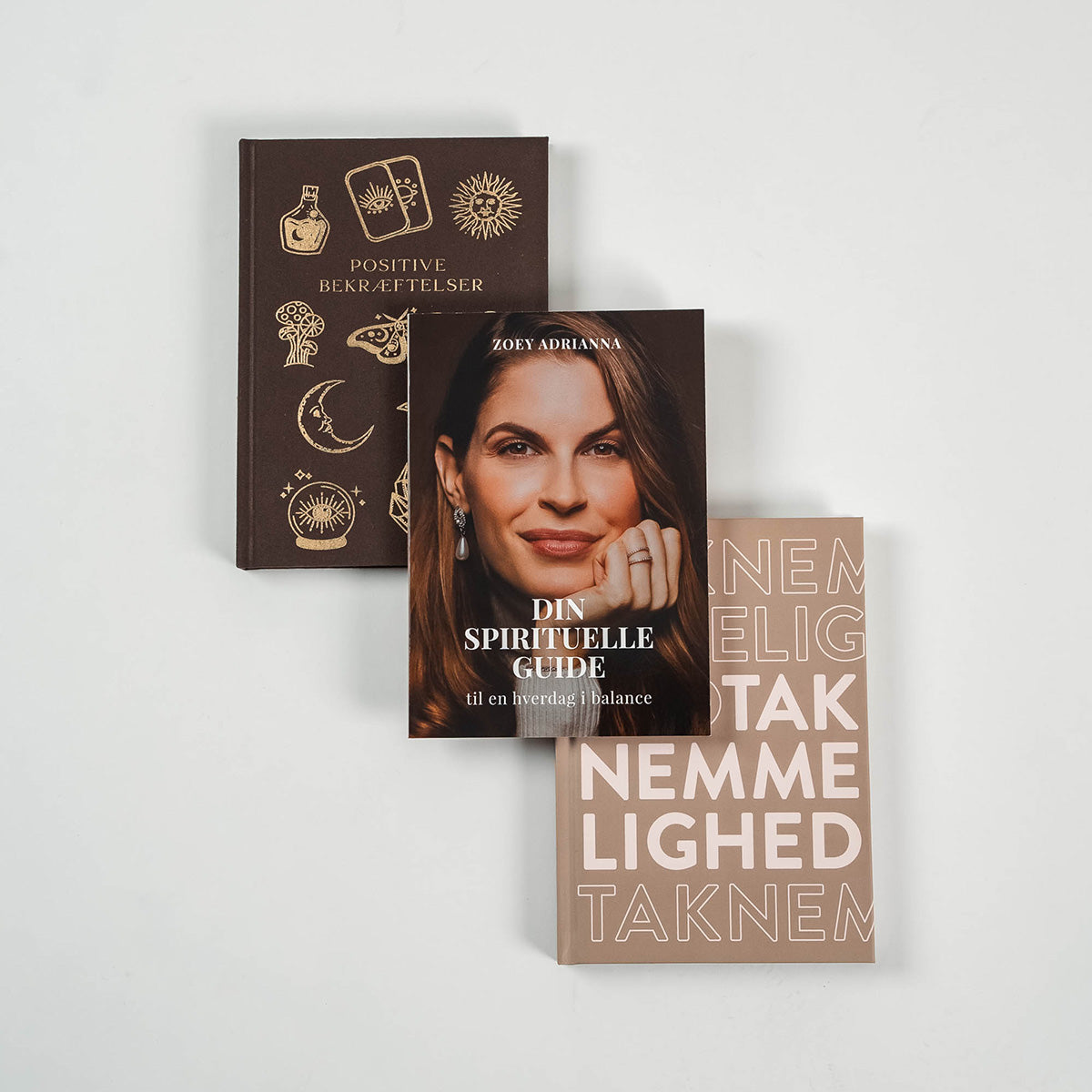 Tre spirituelle bøger i en stabel på et lyst bord den øverste bog er mærket Din Spirituelle Guide til en hverdag i balance og viser et billede af en kvinde med lange brune hår og smykker den midterste bog er mærket Positive Bekræftelser med et mørkt omslag dekoreret med symbolske illustrationer den nederste bog er mærket Taknemmelighed med store letlæselige bogstaver i en neutral farve