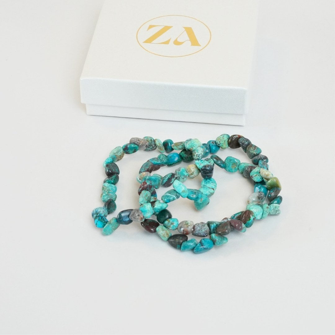 Chrysocolla armbånd i nuancer af turkis, grøn og brun ligger tilfældigt på en hvid overflade sammen med en hvid gaveæske med guldfarvet logo