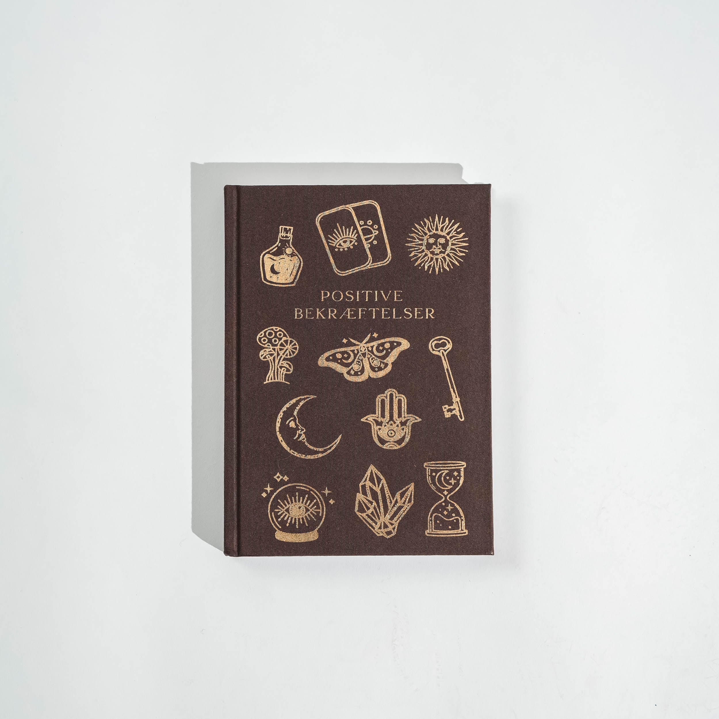 Mørkebrune notesbog med gyldne illustrationer og teksten Positive bekræftelser, prydet med symbolske motiver som en hånd, en sommerfugl, en nøgle, og stjerner