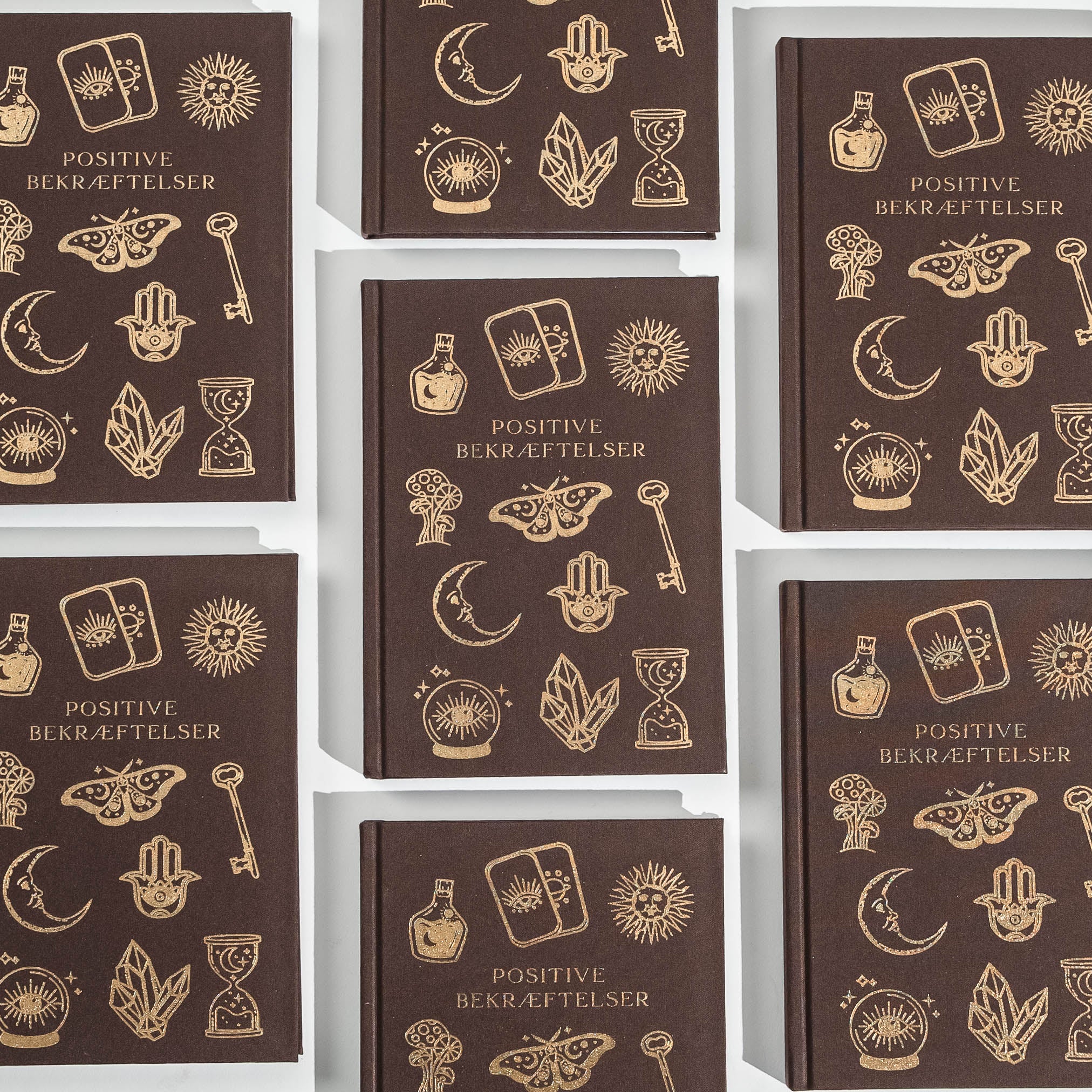 En samling af notesbøger med titlen Positive bekræftelser af Zoey Adrianna. Hver notesbog har en mørk brun omslag prydet med guldtegn og symboler som sol, måne, ur, krystaller og forskellige motiver. Designet er stilfuldt og inspirerende, perfekt til personlige bekræftelser og noter.