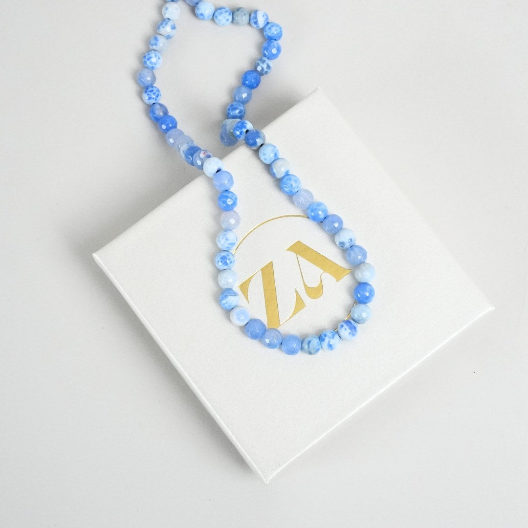PEACEFUL HARMONY halskæde med perler i nuancer af blå på et hvidt smykkeskrin med et gyldent logo
