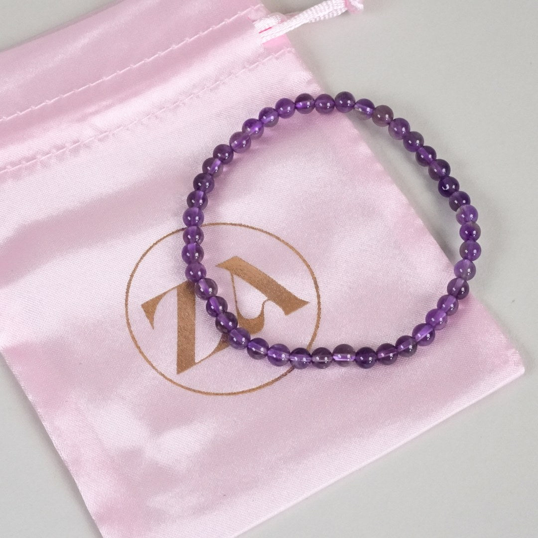 Ametyst armbånd med 4 mm perler præsenteret på en lyserød stofpose med et guldlogo