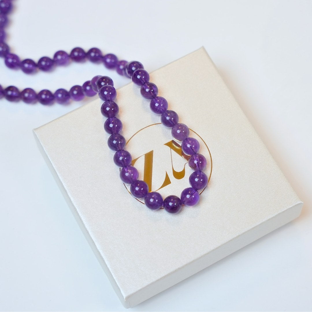Halskæde kaldet HIGH VIBE lavet af lilla ametyst perler præsenteret på en hvid æske med et guld logo