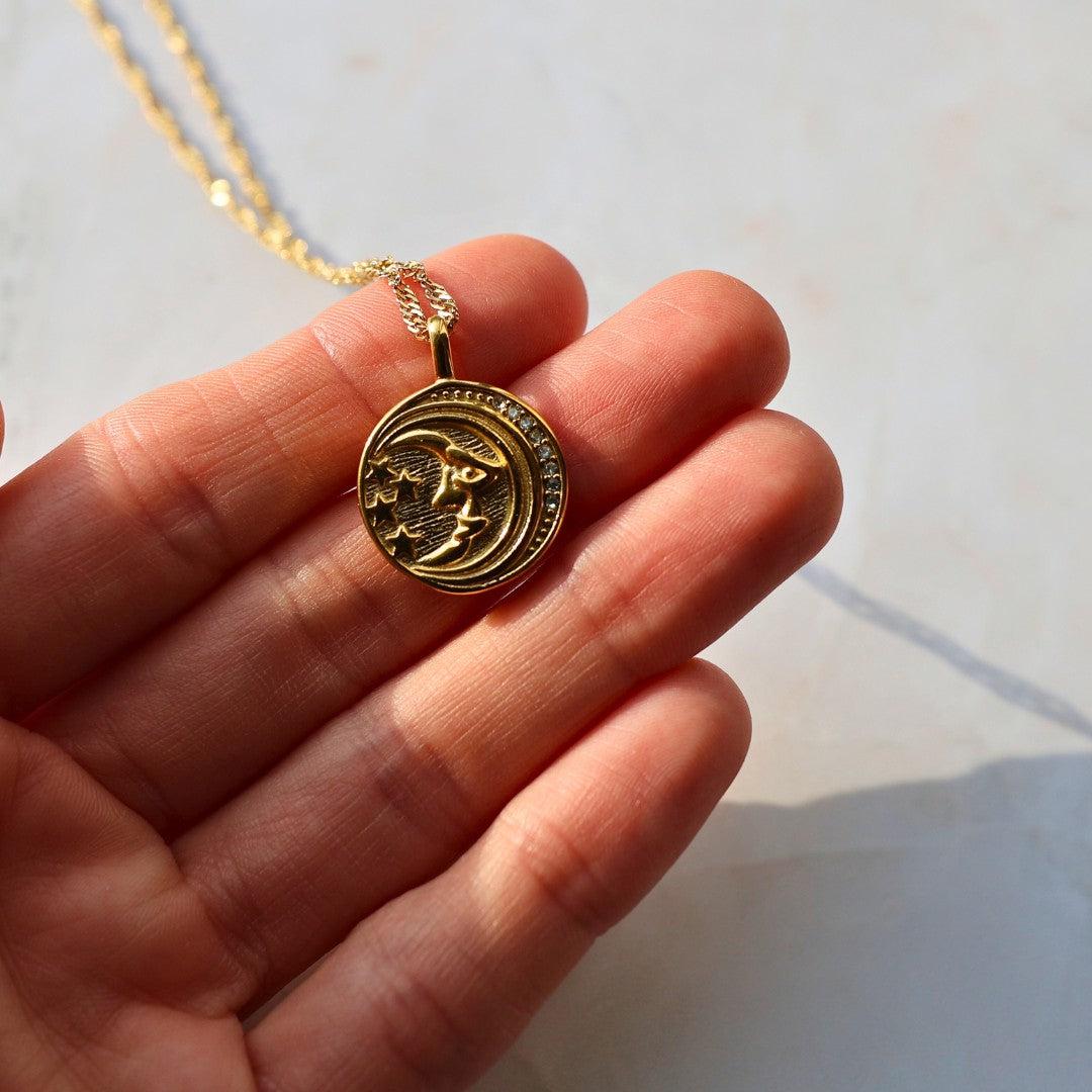 Et guldhalskæde med en cirkulær vedhæng der viser et ansigt med stjerner og måne i et detaljeret design holdt i en hånd