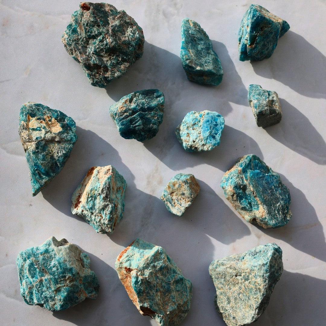 En samling af blå apatitrå mineraler i forskellige størrelser og former, præsenteret på en lys overflade med skygger fra sollys, der tilføjer dybde til billedet.