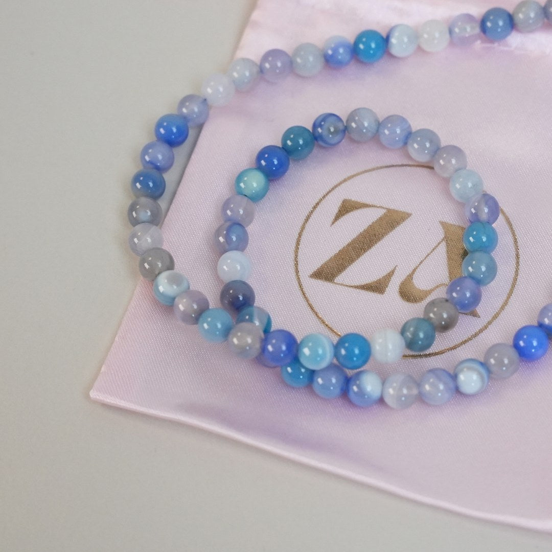 Halskæde i nuancer af blå og grå med runde perler af forskellige størrelser præsenteret på en blød lys pink pose med et gyldent logo