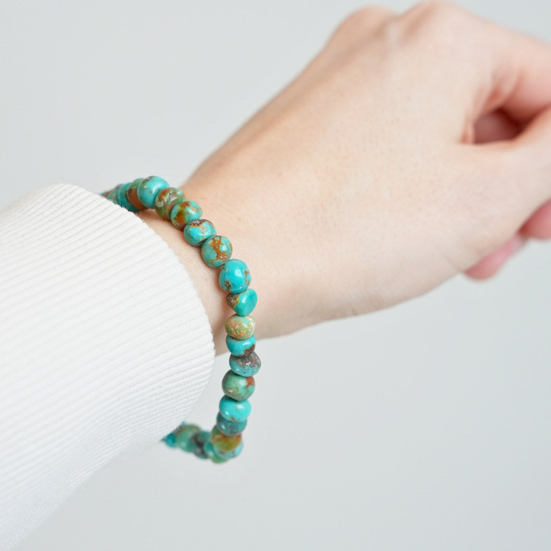 Armbånd i turkise nuancer båret på en hånd med en lys sweater, viser enkelte stykker af glansfuld sten og en rustik finish