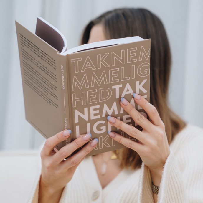 Zoey Adrianna iført en cremefarvet striktrøje sidder afslappet og holder en taknemlighedsdagbog op foran sig. Hendes velplejede negle har en elegant lyseblå kant, og bogen har et stilrent design med ordet "taknemmelighed" gentaget i forskellige skrifttyper. Baggrunden er lys og rolig, hvilket understøtter en stemning af refleksion og selvomsorg.