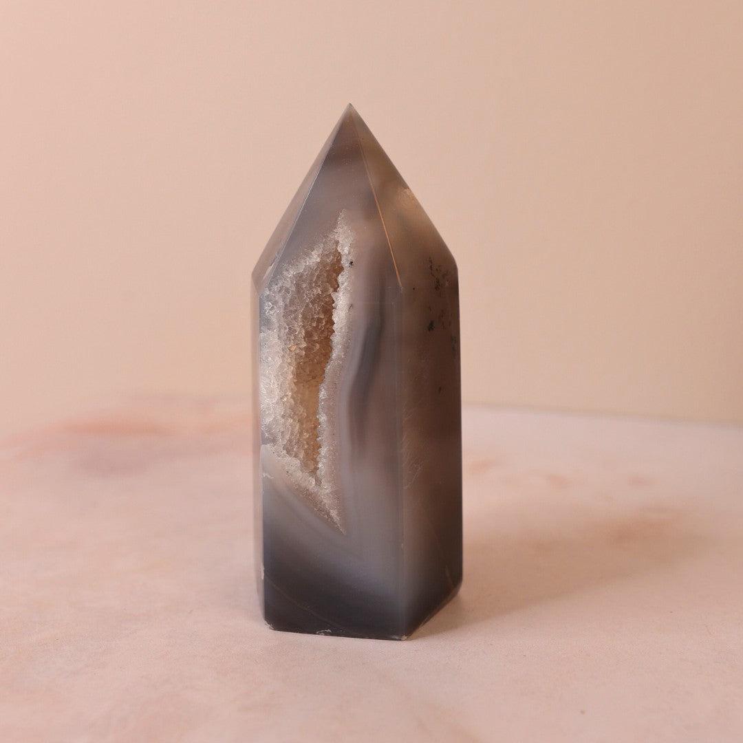 Druzy agat tårn med spids top og en glitrende inderside der viser krystaller i beige og grå nuancer stående på en lys overflade