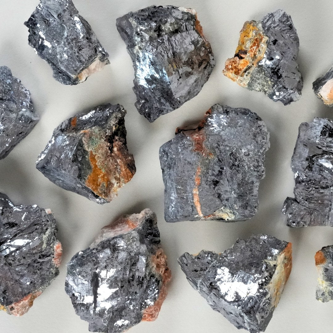 Galenit råstykker i forskellige størrelser og former med sølvgrå overflader og spor af orange og grønne mineraler spredt på en lys baggrund