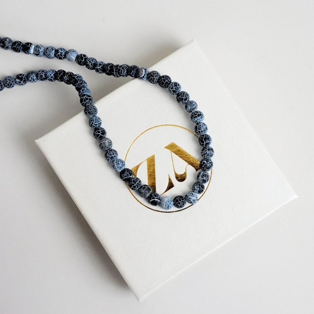 Billede af et halskæde med sorte og blå marmorering perler placeret på en hvid gaveæske med et gyldent logo på toppen