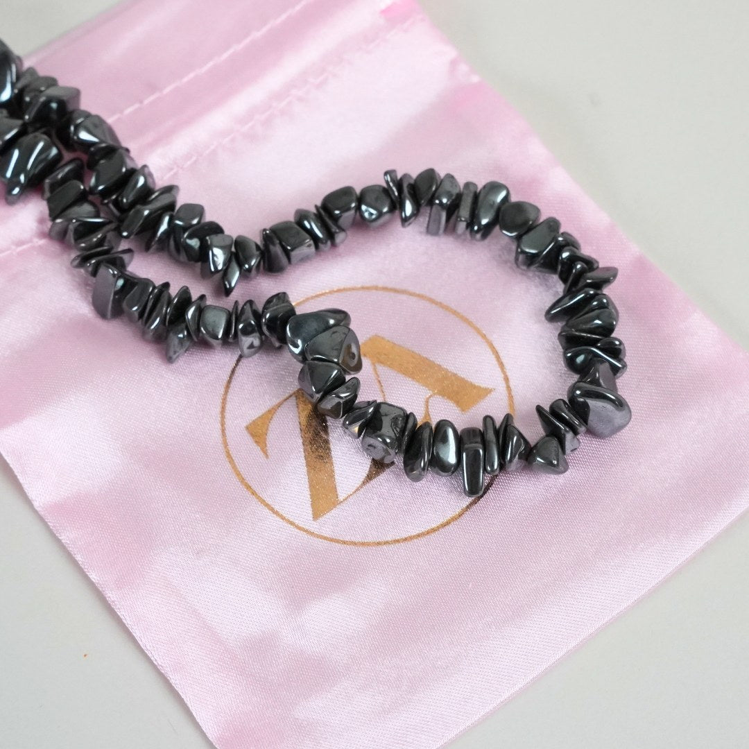 Hæmatit halskæde lagt ud på en lys pink stofpose med et guld logo i midten halskæden er lavet af glinsende hæmatitperler i nuancer af grå og sort med forskellige facetter og former