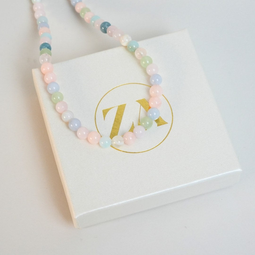 Halskæde med perler i forskellige farver som rosa, grøn, blå og hvid præsenteret på en hvid æske med guld logo