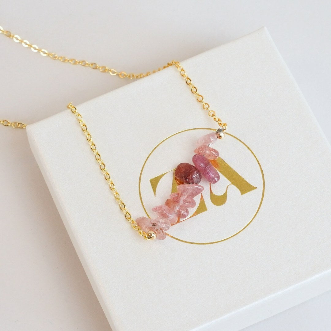 Halskæde med jordbærkvarts perler i forskellige nuancer af pink og lilla, hængende fra en gylden kæde, præsenteret på en hvid æske med logo i guld