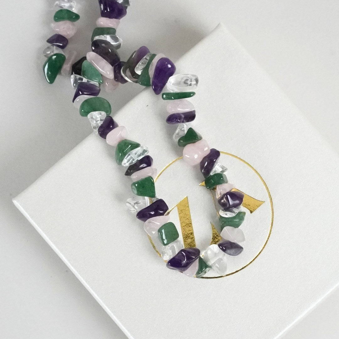 Regnbue halskæde lavet af farverige stykker af natursten som grønne, lilla og klare perler prydet på en hvid gaveæske med et glitrende logo i guld