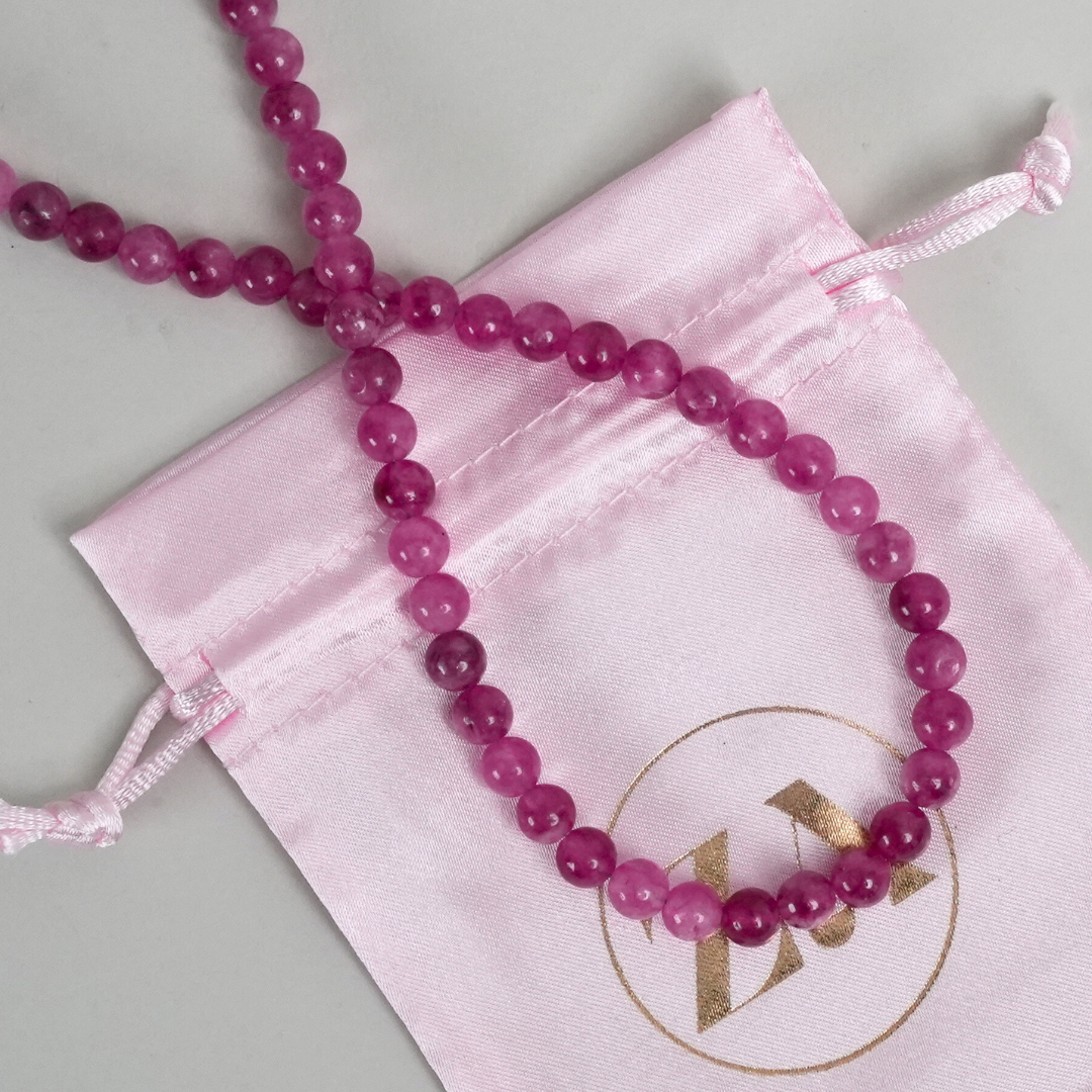 Et smukt halskæde i nuancer af pink perler ligger i en lyserød stofpose med en guldfarvet logo.