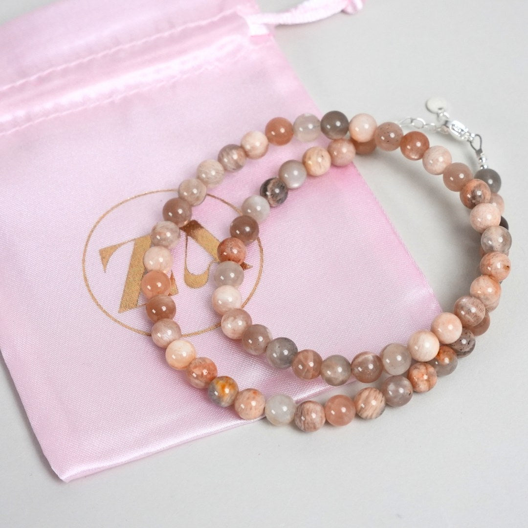 Billede af et smukt SUN halskæde med kugler i nuancer af rosa, beige og grå, præsenteret ved siden af en pink opbevaringspose med en gylden logo detalje