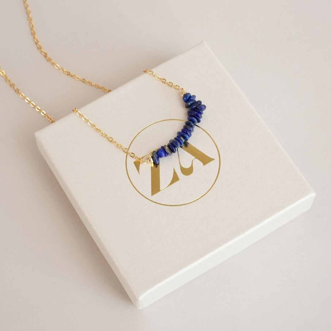 Lapis lazuli halskæde præsenteret på en hvid gaveæske med guldlåg logo designet i midten kæden har blå perler i en bueformet række