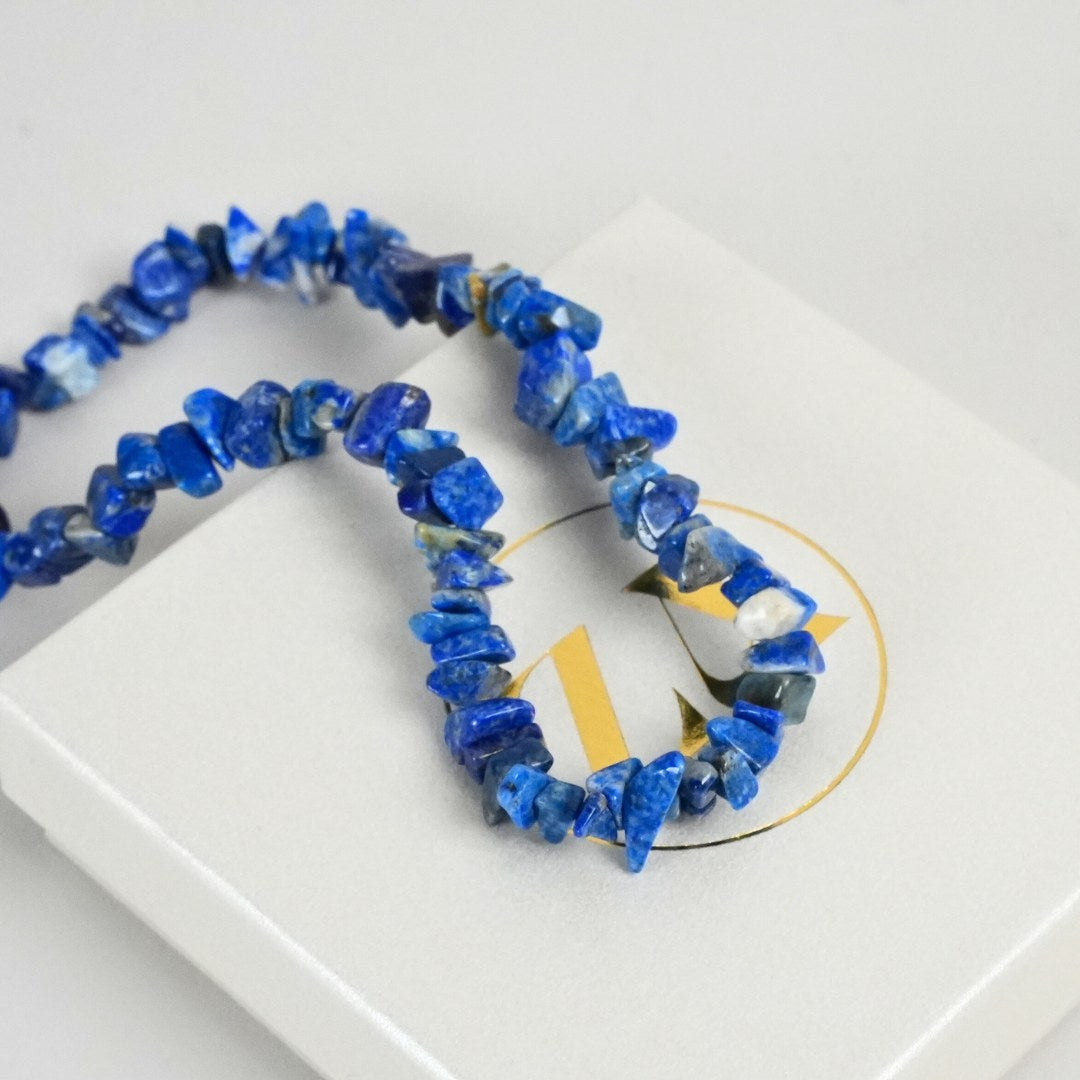 Halskæde lavet af små stykker lapis lazuli i nuancer af blå og gylden, præsenteret på en hvid gaveæske med et dekorativt logo