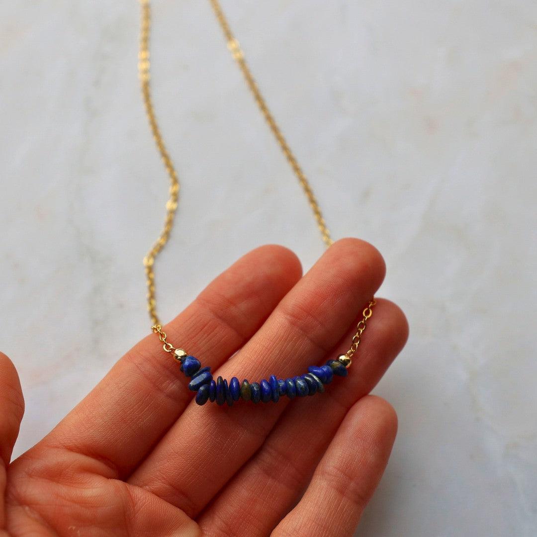 Lapis lazuli halskæde med en gylden kæde præsenteret på en hånd halskæden har en asymmetrisk formation af blå og lilla lapis lazuli perler i forskellige former og størrelser