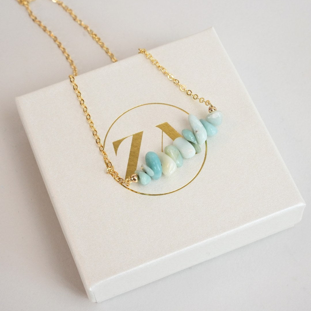Halskæde med larimar perler i forskellige nuancer af blå og grøn på en gylden kæde præsenteret på en hvid gaveæske med logo i guld