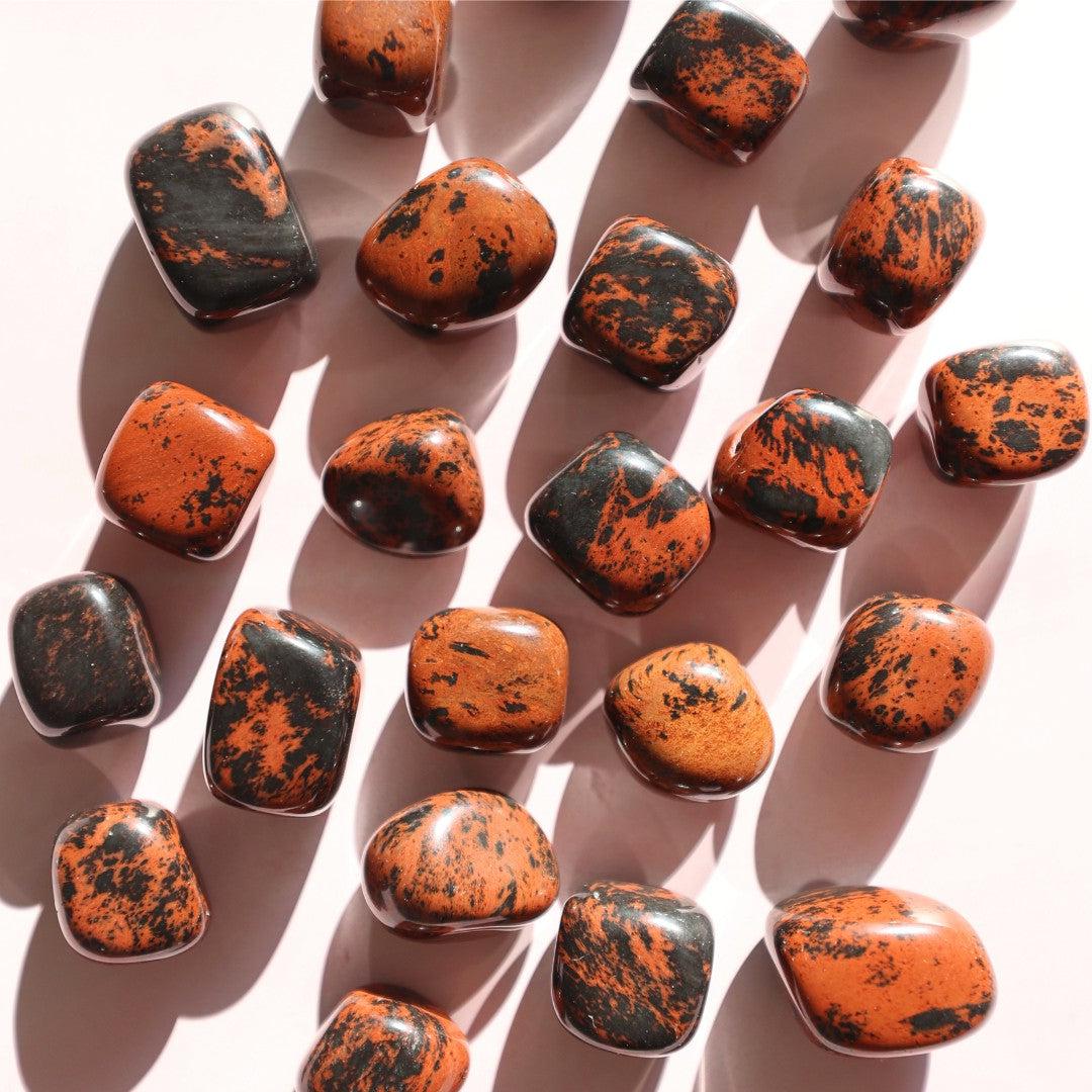 Billede af flere mahogni obsidian sten i forskellige former og størrelser med en glat overflade og en farvekombination af orange og sort, der ligger på en lys baggrund.