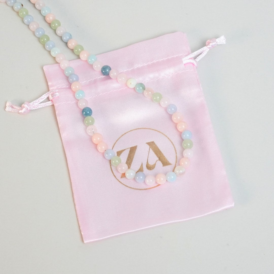 HEALING LOVE halskæde bestående af farverige perler i lyse nuancer af pink, blå og grøn lagt over en lyserød stofpose med guldlogo