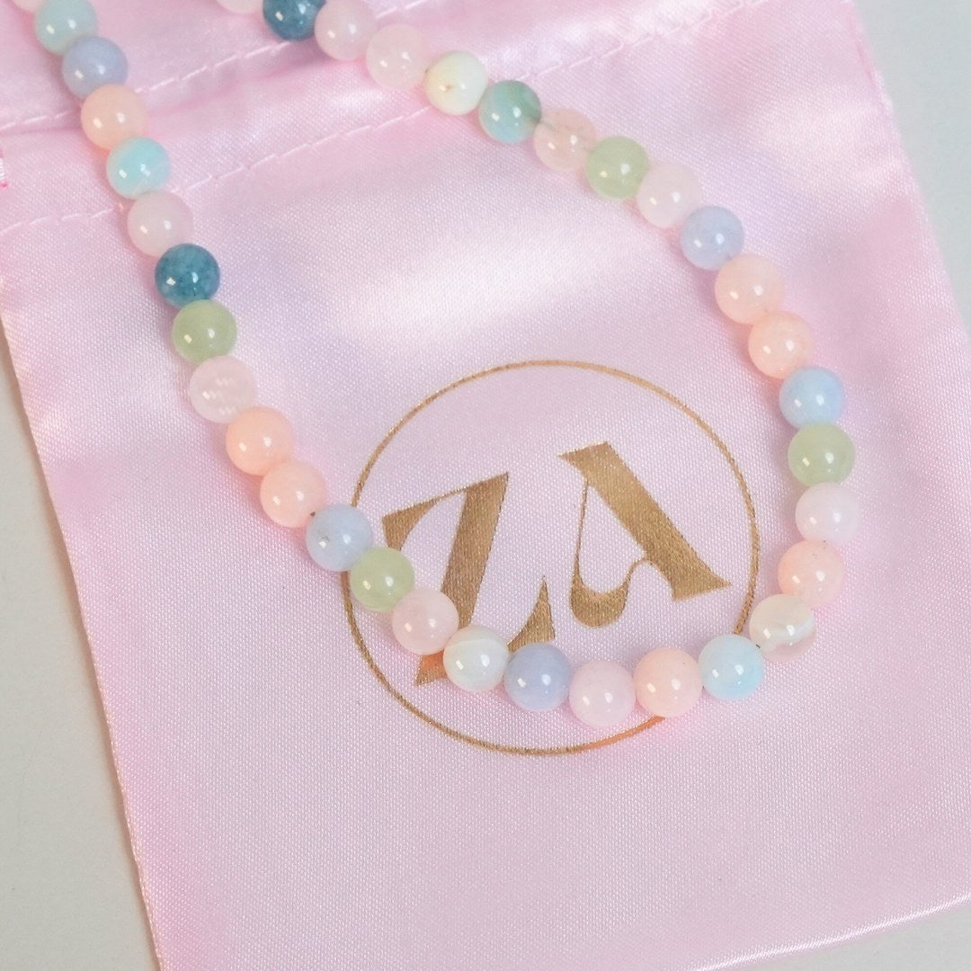 HEALING LOVE halskæde bestående af farverige perler i pasteltoner lagt på en lyserød satinpose med et guldlogo