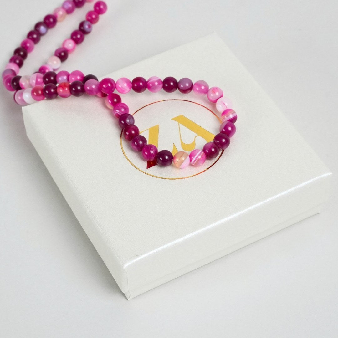 Billede af BALANCE halskæde med perler i nuancer af pink og lilla liggende på en hvid gaveæske med en gylden logo på toppen
