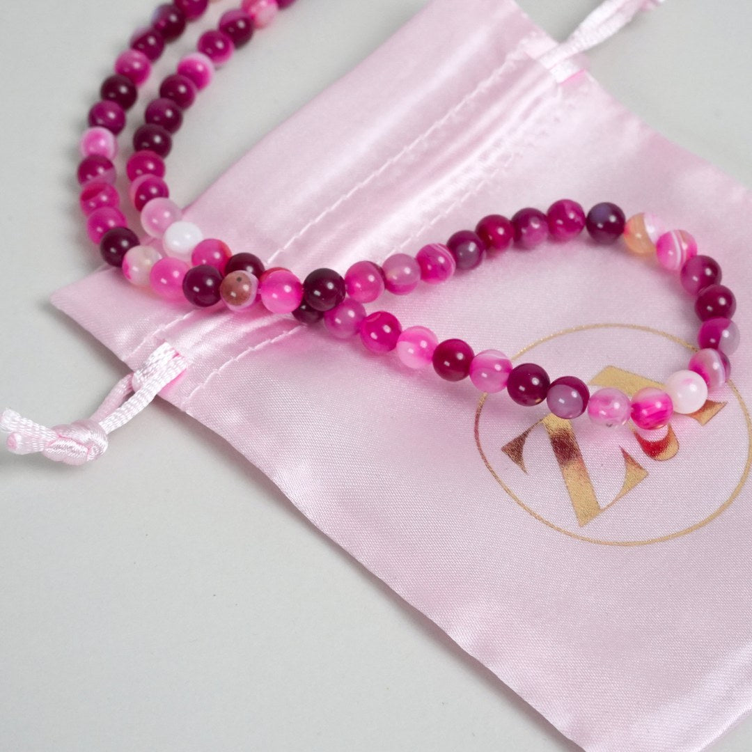 BALANCE halskæde med farverige perler i nuancer af pink og lilla lagt over en lyserød satinpose med logo