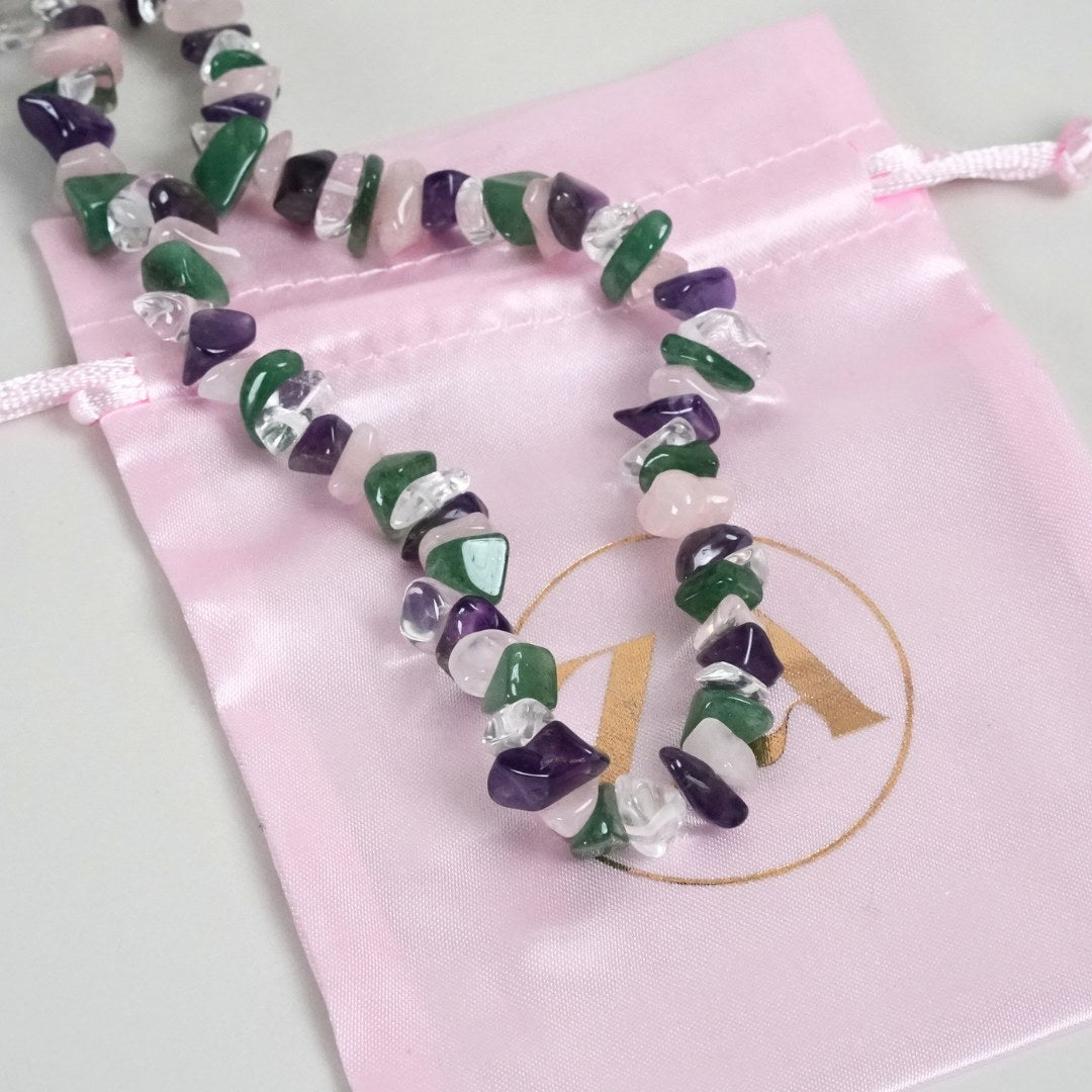 Regnbue halskæde lavet af farverige edelstene som violet, grøn, rosa og gennemsigtig kvarts ligger på en lyserød silkepose med et logo i guld.