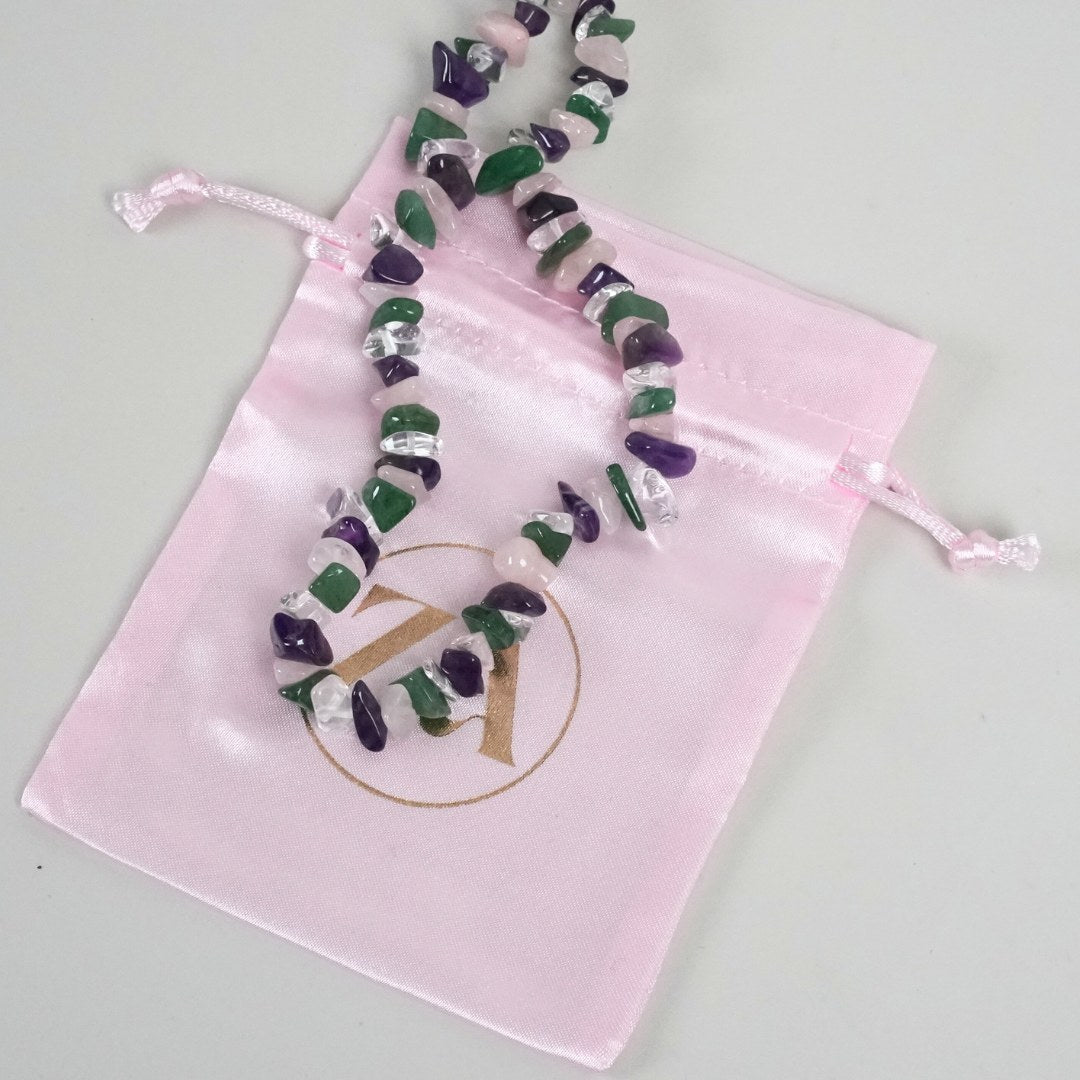 Regnbue halskæde lavet af forskellige halvædelsten i nuancer af grønt, lilla og klart med en pink pose med logo i guld på en lys overflade