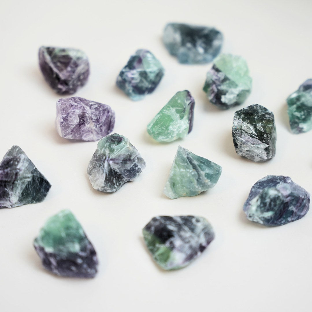 Billede af flere små krystalstykker af regnbue fluorite i nuancer af grøn, lilla og blå der er spredt ud over en lys baggrund