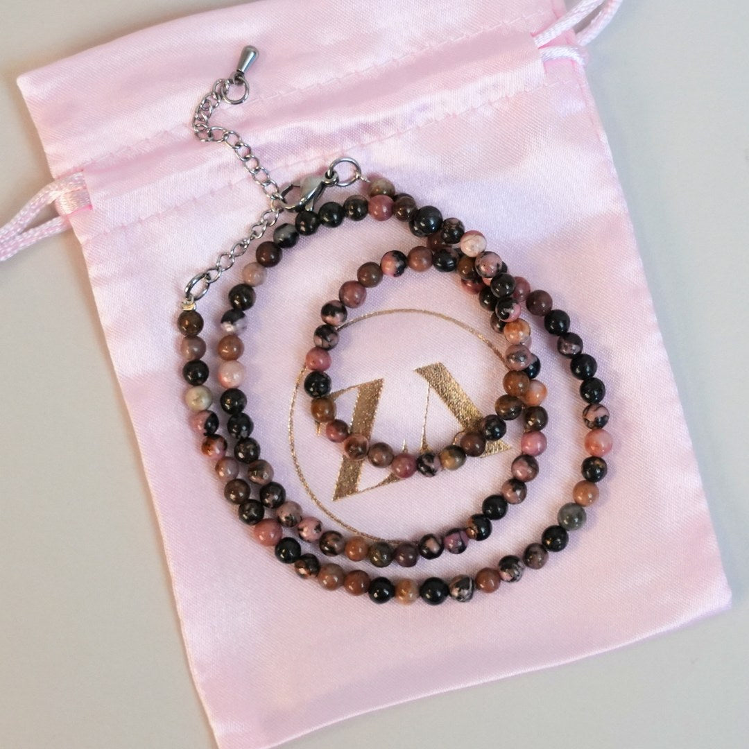HEART halskæde præsenterer en elegant kombination af perler i nuancer af rosa, brun og sort anbragt på et blødt lyserødt stofpose med en guldbelagt logo.