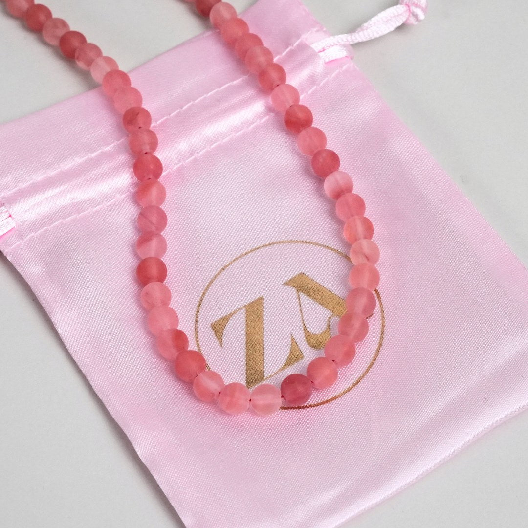 SELF-LOVE halskæde med perler i forskellige nuancer af pink præsenteret på en satinpose i lyserød med guld-logo