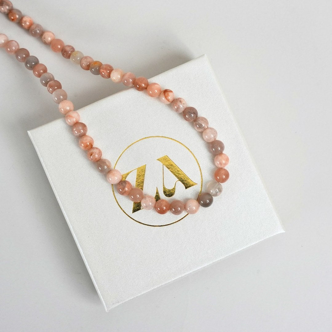 Solen halskæde lavet af smukke rosa og nude perler ligger på en hvid emballage med guld logo