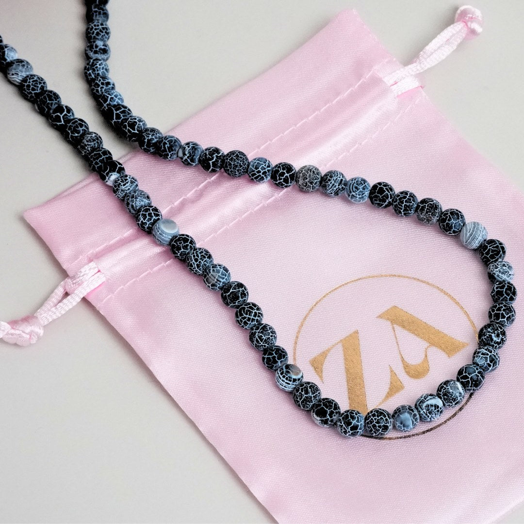 Halskæde lavet af sorte og blå perler med et unikt mønster på en rosa satinpose med et logo i guld.