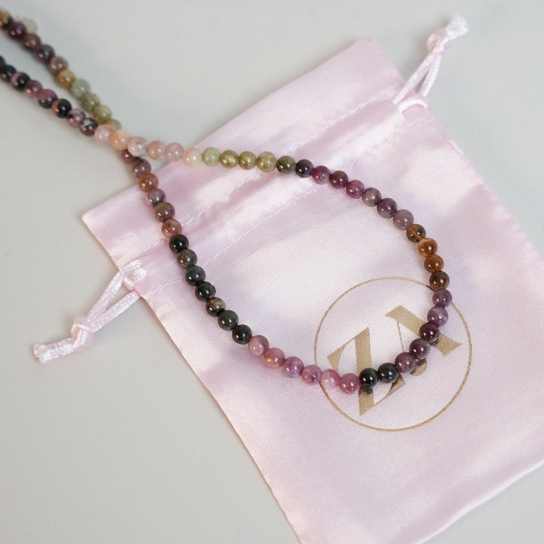 Wisdom halskæde bestående af små, farverige perler med nuancer af pink, grøn, lilla og sort liggende på en lyserød stofpose med et logo