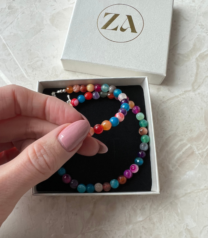 En hånd holder et farverigt HAPPY halskæde i en smuk gaveæske med logoet ZA synligt på låget halskædet består af flere lyse perler i forskellige nuancer og ligger blødt på en sort baggrund inde i æsken
