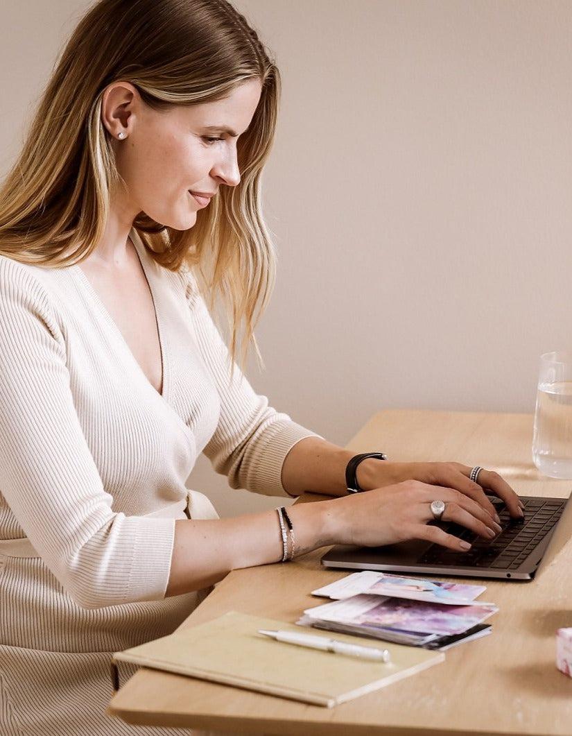 Kvinde med langt hår sidder ved et træbord og arbejder på en bærbar computer hun bærer en lys striktrøje og flere armbånd samt ringe på hænderne der ligger billeder og en notesblok samt en pen på bordet og et glas vand ved siden af