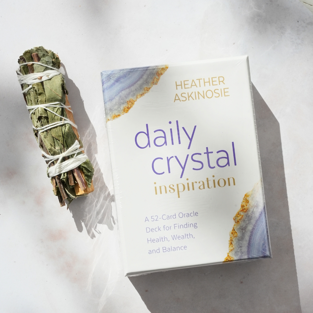 Forpakningen af Daily Crystal Inspiration orakelkort med en beskrivelse af produktet der fremhæver sundhed, rigdom og balance ligger ved siden af et bundt tørret urt.